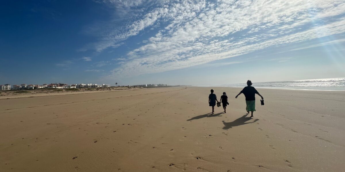 Three silhouetted figures walking on expansive sandy beach in Assilah, Morocco, with dramatic cloudy sky above and coastal buildings visible in distance