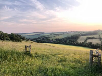 Rolling green hills and farmland in England with wooden fence gates in foreground, scattered village below, and soft pink sunset sky creating peaceful rural landscape