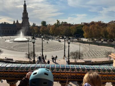 Two children overlooking the magnificent Plaza de España in Sevilla from an ornate ceramic-tiled balustrade, with the iconic tower and fountain visible under dramatic autumn skies