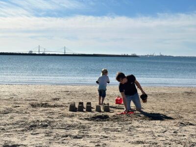 Father and child building sandcastles on sandy beach in El Puerto de Santa María, Spain, with blue water and distant bridge visible under cloudy sky